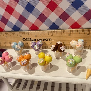 May include: Miniature ice cream cones and scoops in various colors, including pink, orange, yellow, green, and brown, are displayed in clear plastic cups. Each scoop is adorned with colorful sprinkles. A single ice cream cone sits on a white table.