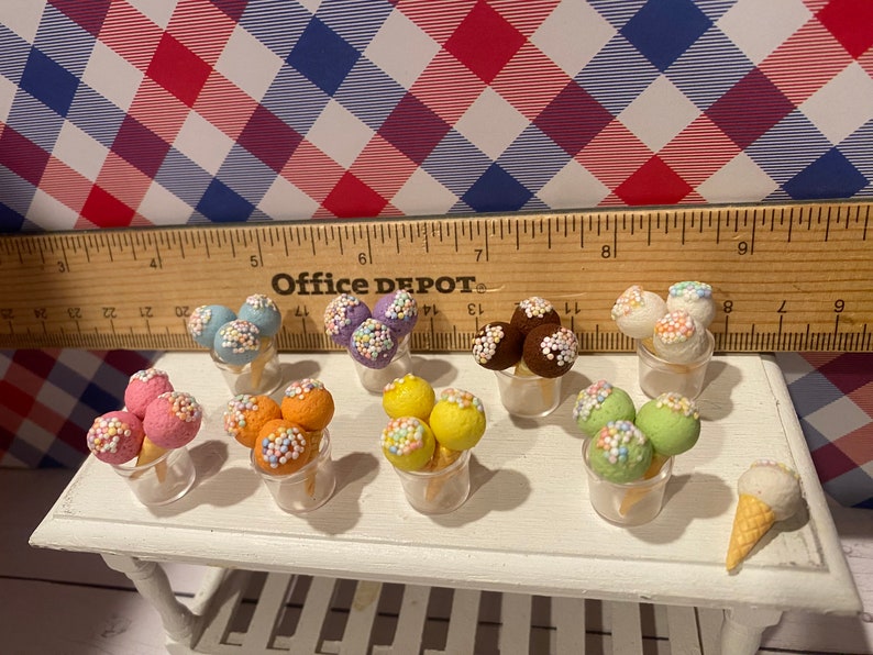 May include: Miniature ice cream cones and scoops in various colors, including pink, orange, yellow, green, and brown, are displayed in clear plastic cups. Each scoop is adorned with colorful sprinkles. A single ice cream cone sits on a white table.