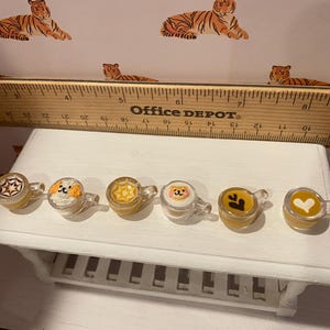 Pode incluir: Seis canecas transparentes em miniatura com v&aacute;rios desenhos, incluindo uma estrela, uma cara de c&atilde;o e um cora&ccedil;&atilde;o. As canecas s&atilde;o preenchidas com um l&iacute;quido amarelo e est&atilde;o sobre uma superf&iacute;cie branca. Uma r&eacute;gua de madeira est&aacute; no fundo.