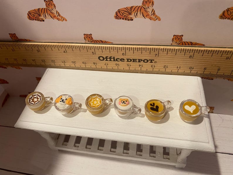 Pode incluir: Seis canecas transparentes em miniatura com v&aacute;rios desenhos, incluindo uma estrela, uma cara de c&atilde;o e um cora&ccedil;&atilde;o. As canecas s&atilde;o preenchidas com um l&iacute;quido amarelo e est&atilde;o sobre uma superf&iacute;cie branca. Uma r&eacute;gua de madeira est&aacute; no fundo.