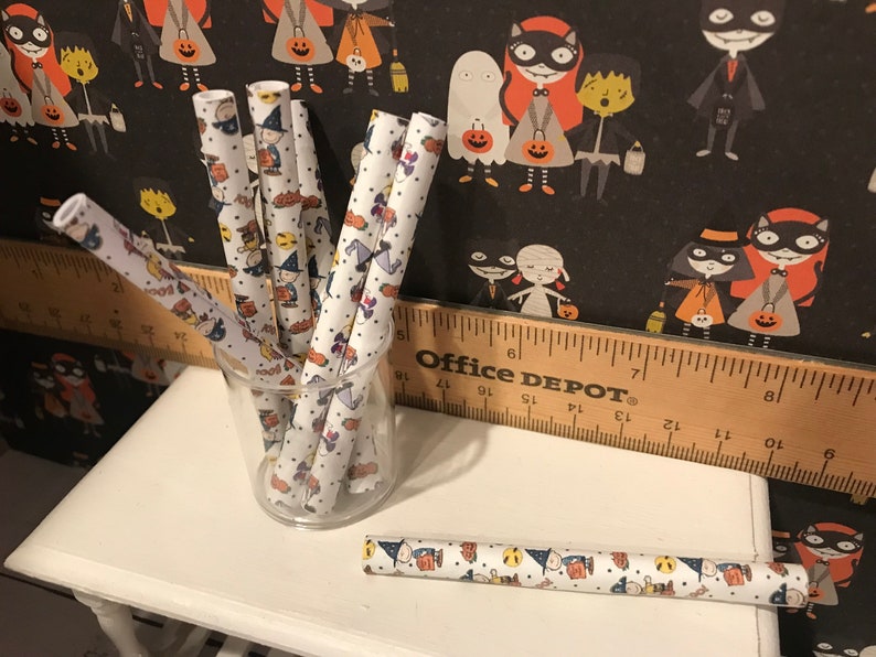 Puede incluir: Un juego de pajitas de papel blancas con un estampado de Halloween con personajes negros y naranjas. Las pajitas est&aacute;n en un vaso transparente sobre una mesa blanca con una regla de madera que dice "Office Depot".