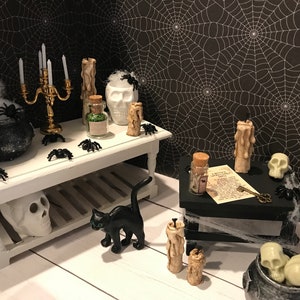 Puede incluir: Una escena de Halloween en miniatura con un gato negro, un caldero, una calavera, velas y frascos sobre una mesa blanca y una mesa negra con un patr&oacute;n de telara&ntilde;a.