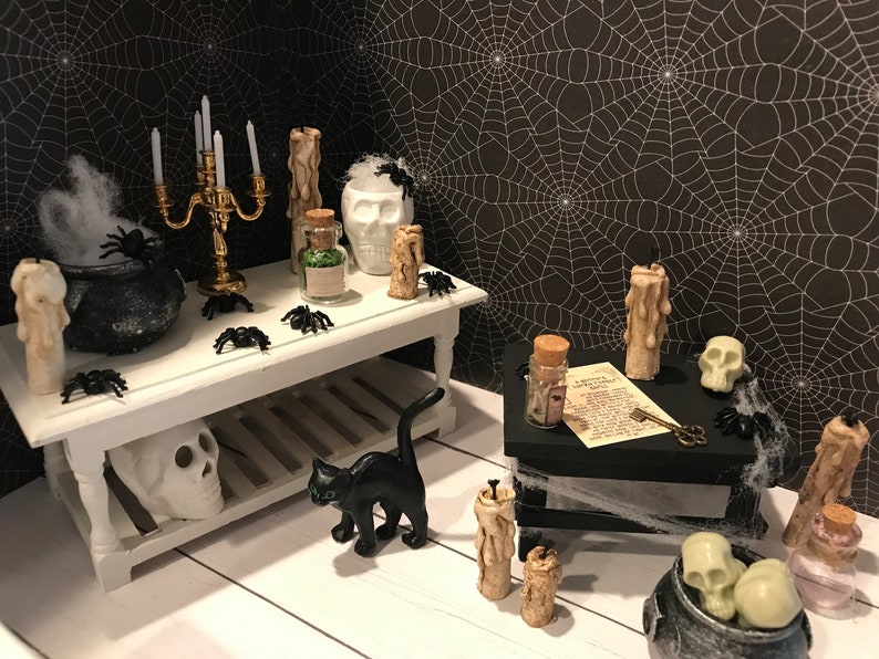 Puede incluir: Una escena de Halloween en miniatura con un gato negro, un caldero, una calavera, velas y frascos sobre una mesa blanca y una mesa negra con un patr&oacute;n de telara&ntilde;a.