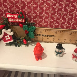 May include: Miniature Christmas decorations on a white table. Includes a wreath with "Merry Christmas" text, a red Santa Claus figure, and two snowman mugs. The background features a red and white snowflake pattern.