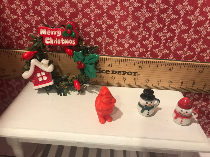 May include: Miniature Christmas decorations on a white table. Includes a wreath with "Merry Christmas" text, a red Santa Claus figure, and two snowman mugs. The background features a red and white snowflake pattern.