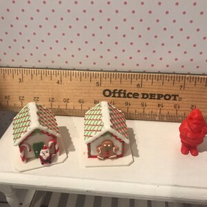 May include: Three miniature gingerbread houses decorated with red and green icing, one with a gingerbread man, one with a Santa Claus figurine, and one with candy canes. A red Santa Claus figurine is also included.