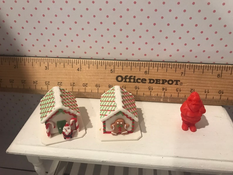 May include: Three miniature gingerbread houses decorated with red and green icing, one with a gingerbread man, one with a Santa Claus figurine, and one with candy canes. A red Santa Claus figurine is also included.