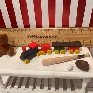 May include: A collection of miniature toys including a brown teddy bear, a blue and red toy train, a baseball bat, a baseball, and a brown baseball glove. The toys are arranged on a white table.