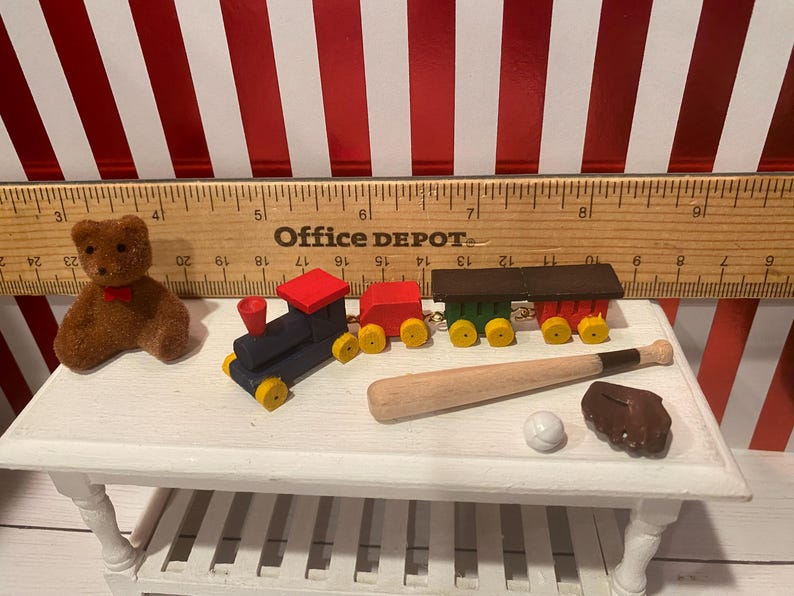 May include: A collection of miniature toys including a brown teddy bear, a blue and red toy train, a baseball bat, a baseball, and a brown baseball glove. The toys are arranged on a white table.