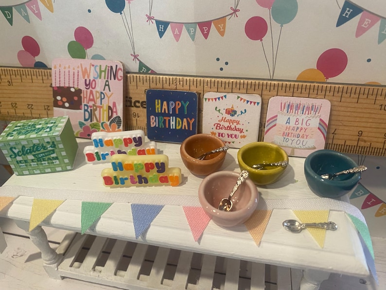 May include: Miniature birthday-themed decorations, including colorful cards, bowls with spoons, and a banner. The cards feature birthday messages. There is also a miniature ice cream box and a cake-shaped decoration.