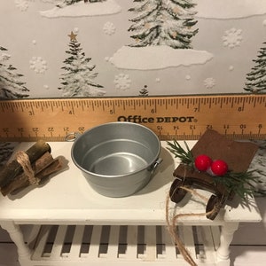 May include: A miniature Christmas scene with a silver metal bucket, a bundle of wooden logs tied with twine, and a wooden sled with red berries and green sprigs. The scene is set on a white wooden table with a ruler in the background.