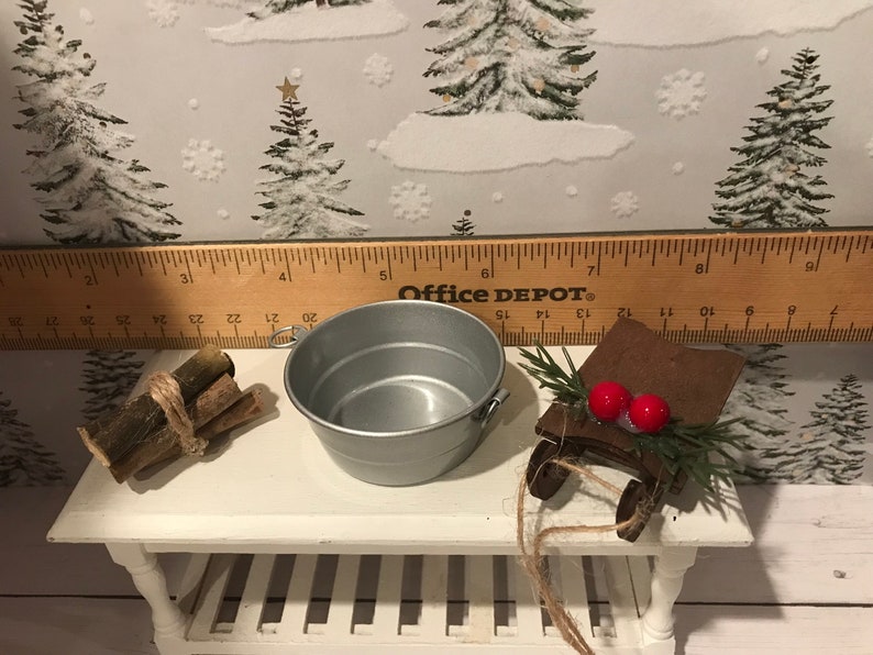May include: A miniature Christmas scene with a silver metal bucket, a bundle of wooden logs tied with twine, and a wooden sled with red berries and green sprigs. The scene is set on a white wooden table with a ruler in the background.