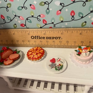 May include: A white miniature table with four miniature Christmas desserts. The desserts include a red and white glazed ham, a red and white pie, a white cake with a snowman on top, and a white cake with a red heart and a snowman on top.