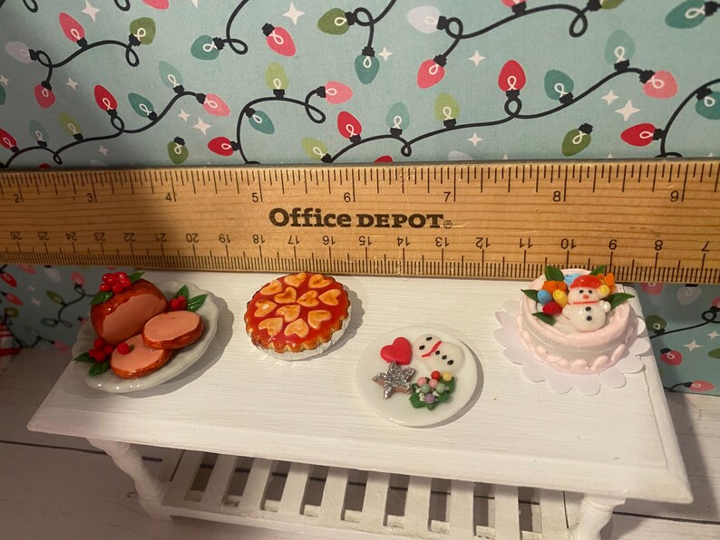 May include: A white miniature table with four miniature Christmas desserts. The desserts include a red and white glazed ham, a red and white pie, a white cake with a snowman on top, and a white cake with a red heart and a snowman on top.