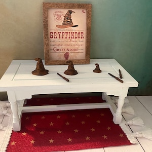 May include: A white table with a red rug with gold stars. There are four brown sorting hats and two wooden wands on the table. A framed poster with the words "You might belong in Gryffindor" is behind the table.