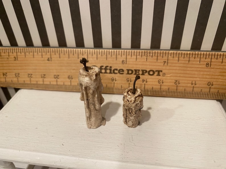 May include: Two small, off-white, textured candles with dark wicks. One candle is taller than the other. A wooden ruler with black markings and the text "office DEPOT" is in the background.