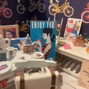 May include: A miniature tea party scene featuring a white tea set, a teapot shaped like Big Ben, and a vintage postcard. The scene is set on a white table with a brown suitcase and a framed picture of Queen Elizabeth II. The background features a blue wall with a repeating pattern of bicycles and the words "Enjoy Tea!" on a blue and white poster.