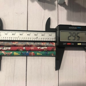Puede incluir: Un calibre digital negro que mide tres pajitas de papel de colores con tem&aacute;tica navide&ntilde;a. El calibre muestra 2,95 pulgadas.