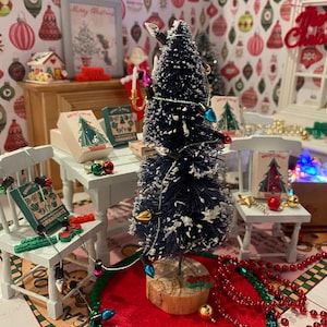May include: A miniature Christmas scene with a small white table, two chairs, and a flocked artificial Christmas tree with white lights. The tree is decorated with red and white ornaments and is surrounded by red and white beaded garland. The table is decorated with small boxes and a book that says "Christmas Ornaments".