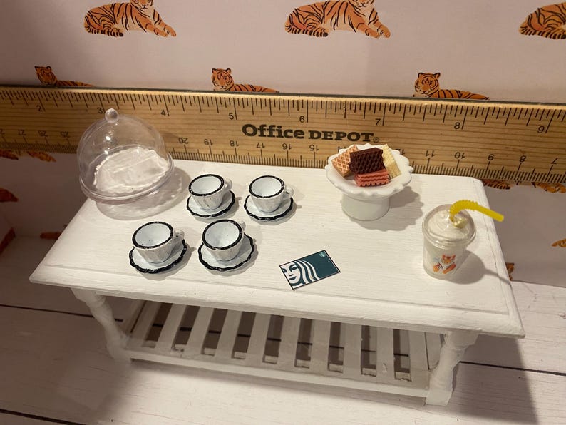 Pode incluir: Mesa branca em miniatura com um conjunto de x&iacute;caras e pires, um prato de sobremesa com biscoitos e um cart&atilde;o-presente Starbucks. Uma c&uacute;pula transparente e uma bebida com canudo amarelo tamb&eacute;m est&atilde;o na mesa. O fundo apresenta uma r&eacute;gua e estampa de tigre.