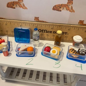 Pode incluir: V&aacute;rios alimentos em miniatura em recipientes transparentes, incluindo frutas, sandu&iacute;ches e doces, s&atilde;o dispostos sobre uma mesa branca. H&aacute; tamb&eacute;m uma pequena garrafa de &aacute;gua, uma garrafa de xarope e uma x&iacute;cara de caf&eacute; Starbucks. Uma r&eacute;gua e papel de parede com tema de tigre est&atilde;o no fundo.