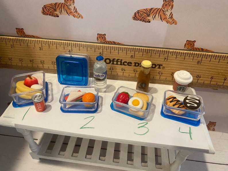 Pode incluir: V&aacute;rios alimentos em miniatura em recipientes transparentes, incluindo frutas, sandu&iacute;ches e doces, s&atilde;o dispostos sobre uma mesa branca. H&aacute; tamb&eacute;m uma pequena garrafa de &aacute;gua, uma garrafa de xarope e uma x&iacute;cara de caf&eacute; Starbucks. Uma r&eacute;gua e papel de parede com tema de tigre est&atilde;o no fundo.