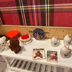Puede incluir: Miniaturas con tem&aacute;tica de osos de peluche sobre una mesa blanca. Incluye un oso de peluche blanco y marr&oacute;n, un pastel rojo, dos revistas y dos tazas con crema batida. Tambi&eacute;n hay una tapa roja y un plato decorativo.