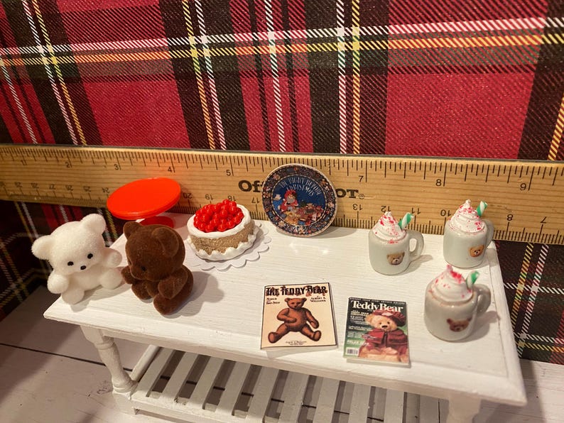 Puede incluir: Miniaturas con tem&aacute;tica de osos de peluche sobre una mesa blanca. Incluye un oso de peluche blanco y marr&oacute;n, un pastel rojo, dos revistas y dos tazas con crema batida. Tambi&eacute;n hay una tapa roja y un plato decorativo.