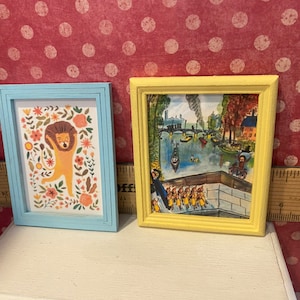 Puede incluir: Dos peque&ntilde;as impresiones art&iacute;sticas enmarcadas. Una presenta una ilustraci&oacute;n de le&oacute;n con acentos florales en un marco azul claro. La otra tiene una escena de paisaje en un marco amarillo. Ambas se exhiben sobre una superficie blanca sobre un fondo rojo con lunares.