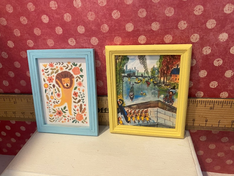 Puede incluir: Dos peque&ntilde;as impresiones art&iacute;sticas enmarcadas. Una presenta una ilustraci&oacute;n de le&oacute;n con acentos florales en un marco azul claro. La otra tiene una escena de paisaje en un marco amarillo. Ambas se exhiben sobre una superficie blanca sobre un fondo rojo con lunares.