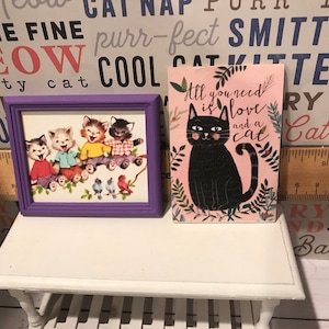 May include: Two framed pictures with cat themes. The first picture is a purple frame with a vintage illustration of four cartoon cats. The second picture is a pink frame with a black cat illustration and the text "All you need is love and a cat".