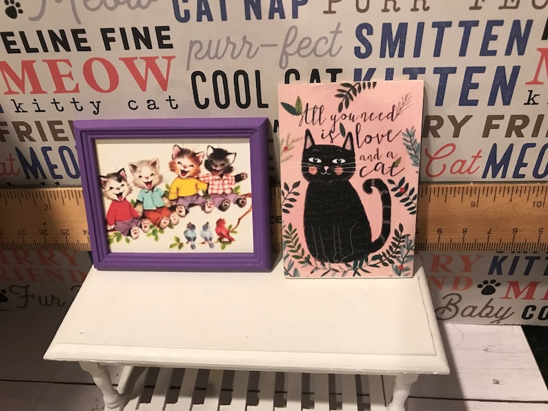 May include: Two framed pictures with cat themes. The first picture is a purple frame with a vintage illustration of four cartoon cats. The second picture is a pink frame with a black cat illustration and the text "All you need is love and a cat".