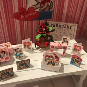Może przedstawiać: Kolekcja kart i dekoracji walentynkowych w stylu vintage na białym stole. Karty przedstawiają romantyczne ilustracje i są eksponowane obok kompozycji kwiatowej i kalendarza na luty. Widoczne jest czerwone serce z napisem "MY VALENTINE".