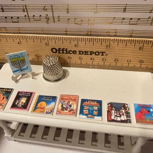 May include: A white table with a silver thimble and eight miniature books about Hanukkah. The books have colorful covers with illustrations of children, animals, and menorahs. The table is on a wooden surface with a ruler that says "Office Depot" on it.
