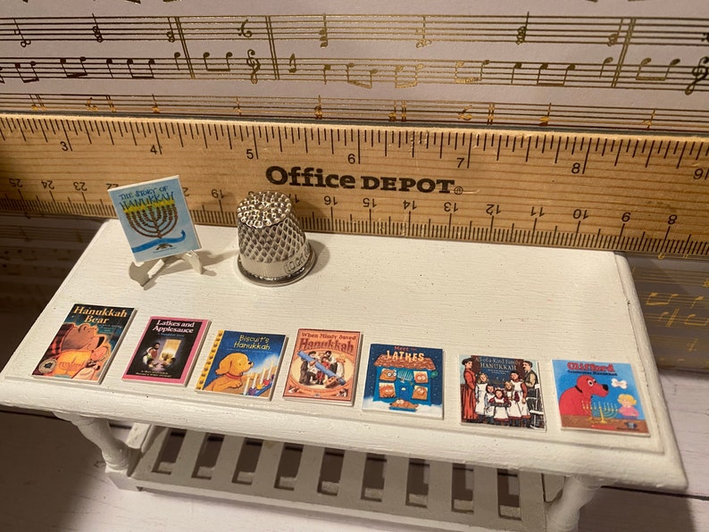 May include: A white table with a silver thimble and eight miniature books about Hanukkah. The books have colorful covers with illustrations of children, animals, and menorahs. The table is on a wooden surface with a ruler that says "Office Depot" on it.