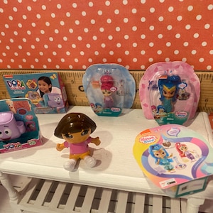 Op de afbeelding: Een verzameling kinderspeelgoed, waaronder Dora the Explorer en Shimmer and Shine beeldjes. Het speelgoed staat op een witte tafel tegen een rode achtergrond met stippen. Sommige speelgoed zit nog in de originele verpakking.