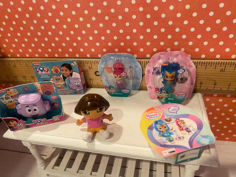 Op de afbeelding: Een verzameling kinderspeelgoed, waaronder Dora the Explorer en Shimmer and Shine beeldjes. Het speelgoed staat op een witte tafel tegen een rode achtergrond met stippen. Sommige speelgoed zit nog in de originele verpakking.