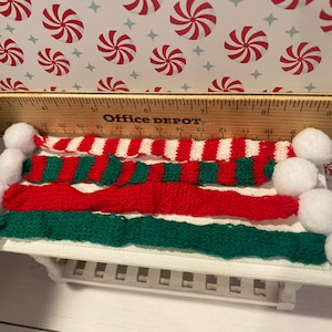 Pode incluir: Um conjunto de tr&ecirc;s cachec&oacute;is de malha em vermelho, branco e verde com pompons brancos nas pontas. Os cachec&oacute;is est&atilde;o dispostos sobre uma mesa branca com uma r&eacute;gua no fundo.