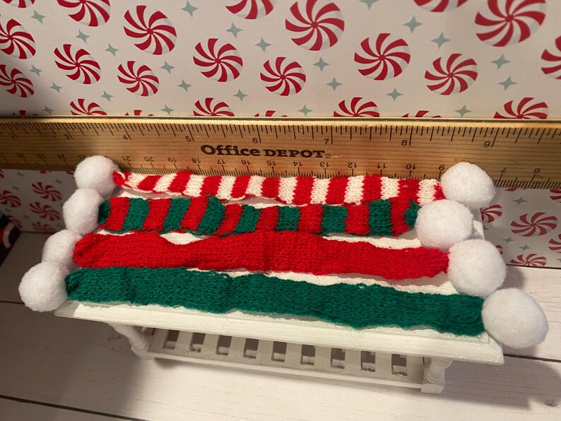Pode incluir: Um conjunto de tr&ecirc;s cachec&oacute;is de malha em vermelho, branco e verde com pompons brancos nas pontas. Os cachec&oacute;is est&atilde;o dispostos sobre uma mesa branca com uma r&eacute;gua no fundo.