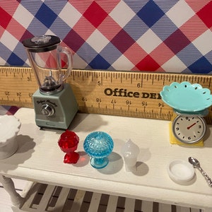 May include: Miniature kitchen set with a blender, scale, and various serving dishes. The blender is light green with a clear container. A small scale with a turquoise plate sits next to a white serving dish. Other items include red and blue glass pieces.