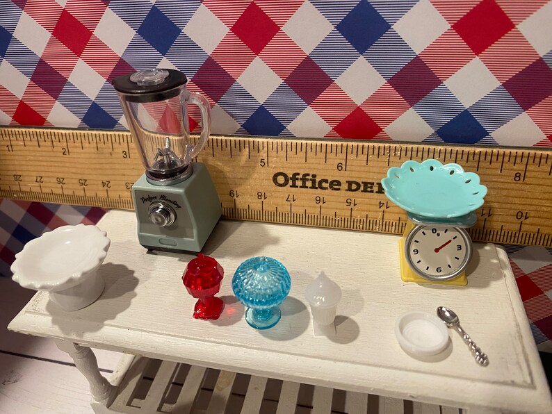 May include: Miniature kitchen set with a blender, scale, and various serving dishes. The blender is light green with a clear container. A small scale with a turquoise plate sits next to a white serving dish. Other items include red and blue glass pieces.