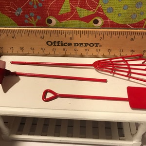 May include: Miniature red garden tools, including a shovel, rake, and hoe, displayed on a white surface. A wooden ruler with "Office DEPOT" printed on it is in the background.