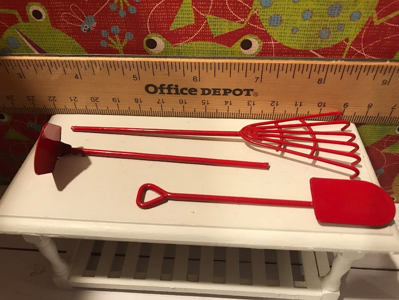 May include: Miniature red garden tools, including a shovel, rake, and hoe, displayed on a white surface. A wooden ruler with "Office DEPOT" printed on it is in the background.