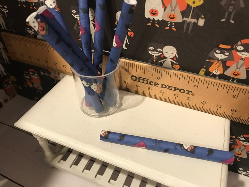 Puede incluir: Un tarro de vidrio lleno de tubos de papel de Halloween azules y blancos. Los tubos est&aacute;n decorados con murci&eacute;lagos, fantasmas y calabazas. El tarro est&aacute; sentado en una mesa blanca con una regla de madera que dice "Office Depot".