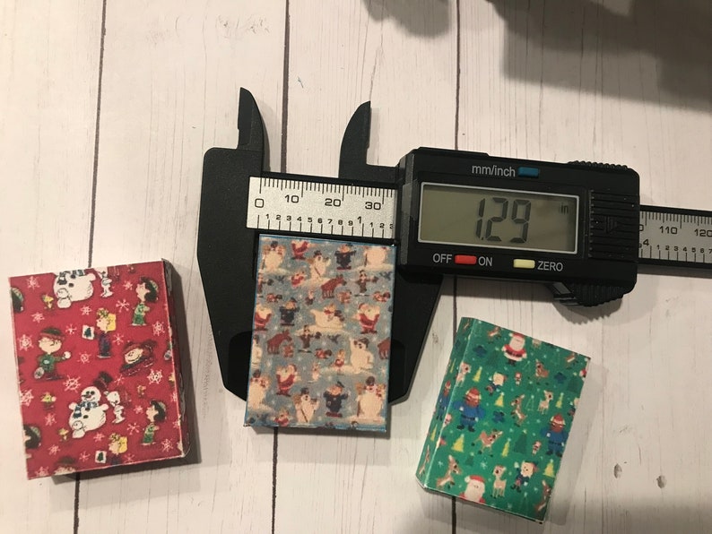 Puede incluir: Tres cajas peque&ntilde;as con diferentes patrones navide&ntilde;os. Las cajas se est&aacute;n midiendo con un calibre, que lee 129 mm. Las cajas tienen aproximadamente 4,5 pulgadas de ancho.