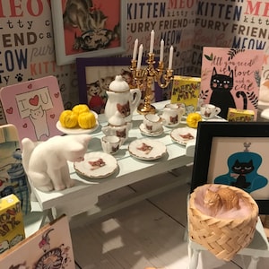 May include: A miniature tea party set with a white teapot, teacups, and saucers decorated with a cat design. The set is arranged on a light blue table with a white cat figurine, yarn balls, and a framed picture of a cat. The table is surrounded by various cat-themed decorations, including signs with the words "Meow" and "Kitty Cat".