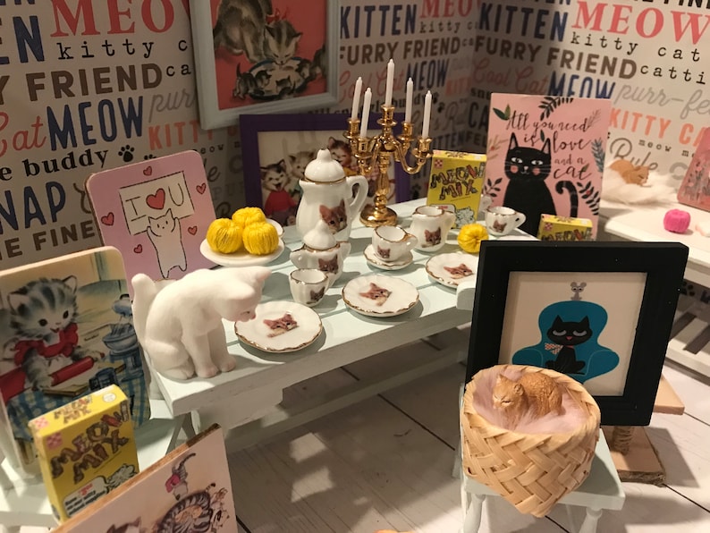 May include: A miniature tea party set with a white teapot, teacups, and saucers decorated with a cat design. The set is arranged on a light blue table with a white cat figurine, yarn balls, and a framed picture of a cat. The table is surrounded by various cat-themed decorations, including signs with the words "Meow" and "Kitty Cat".