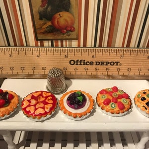 May include: Miniature fruit tarts with various toppings, including berries and kiwi, displayed on a white table. A thimble and a wooden ruler are also present. The ruler is marked in inches.