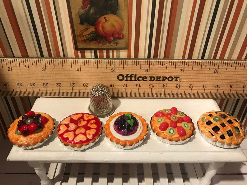 May include: Miniature fruit tarts with various toppings, including berries and kiwi, displayed on a white table. A thimble and a wooden ruler are also present. The ruler is marked in inches.