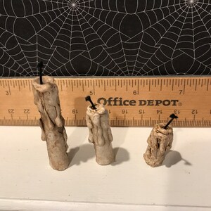 Puede incluir: Tres velas de cera blanca, goteando, con mechas negras. Las velas est&aacute;n dispuestas en fila sobre una superficie blanca. Una regla es visible en el fondo con el texto "Office DEPOT" impreso en ella.